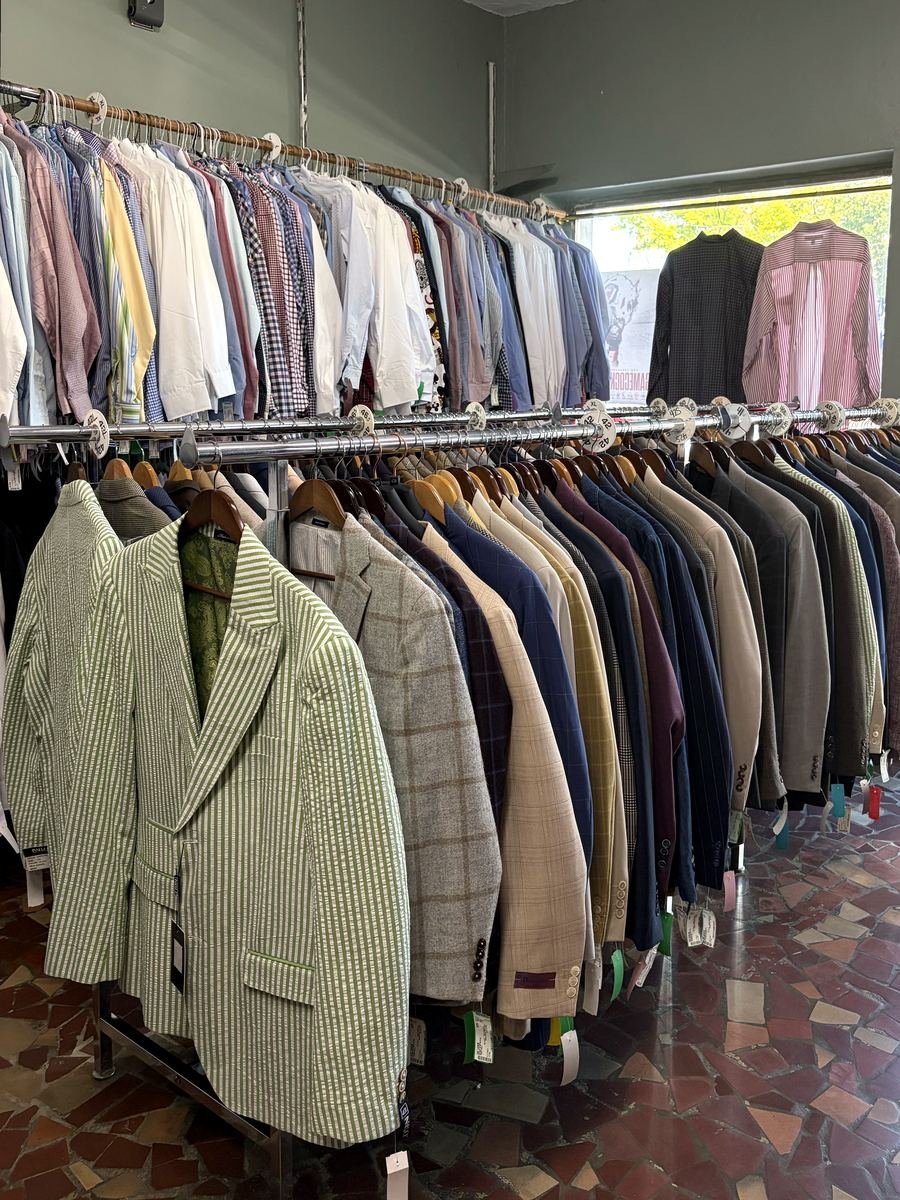 A long rack of sport coats and suit jackets — green seersucker at the front, then tweeds, windowpane checks, plaids and solids — with a full wall of dress shirts behind.