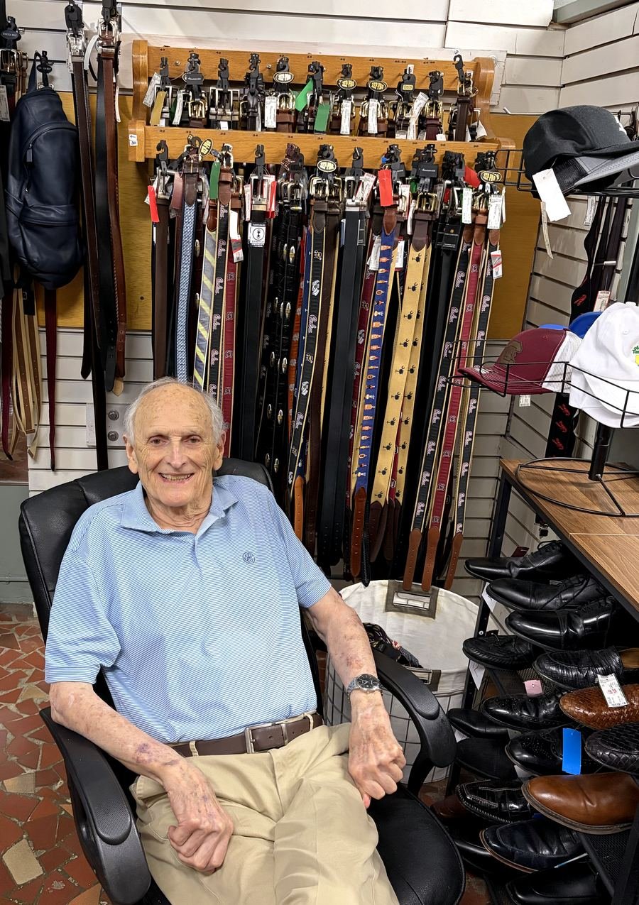 Papa Dean, ninety-five, sitting in the store in a light blue striped polo with a broad smile, a wall of belts behind him.