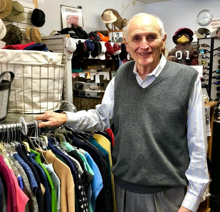 Papa Dean standing with a hand on a rack of sweaters, the store's wall of caps, a framed photo and a USC Gamecock figure behind him.